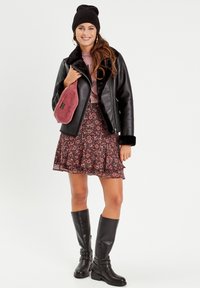 Veste en cuir noir avec col en fausse fourrure, haut rose, jupe à motifs floraux et bottes noires jusqu'aux genoux. Tient une petite pochette rouge.