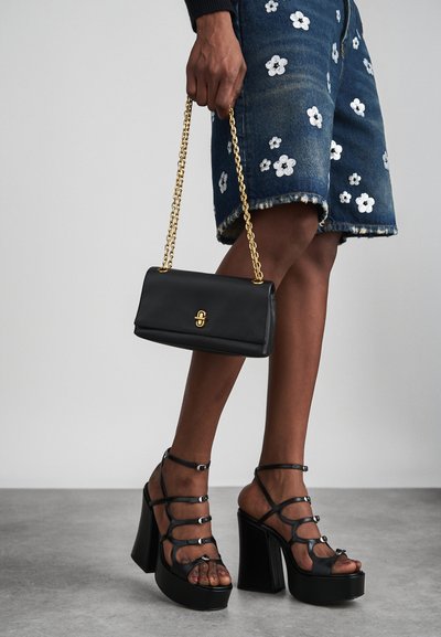 Persoon die een zwarte handtas met gouden ketting vasthoudt, draagt denim shorts met gebloemd borduurwerk en zwarte sandalen met bandjes en plateauzolen.