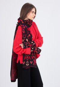 Woman in a red textured sweater wearing a black and red floral patterned scarf and matching fingerless gloves, standing sideways.