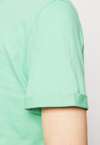 Světl zelené bavlněné tričko s ohrnovaným krátkým rukávem a hladkou texturou, s viditelným švem podél dolního lemu a ramenního švu.