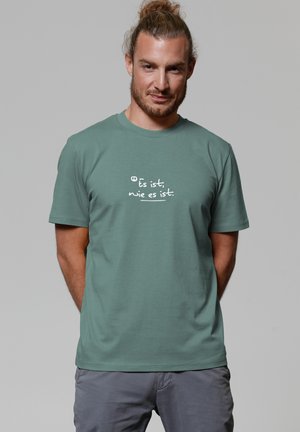 Mann mit hellbraunem Haar im Dutt, der ein grünes T-Shirt mit weißem deutschen Text "Es ist, wie es ist" und grauen Hosen vor einem grauen Hintergrund trägt.