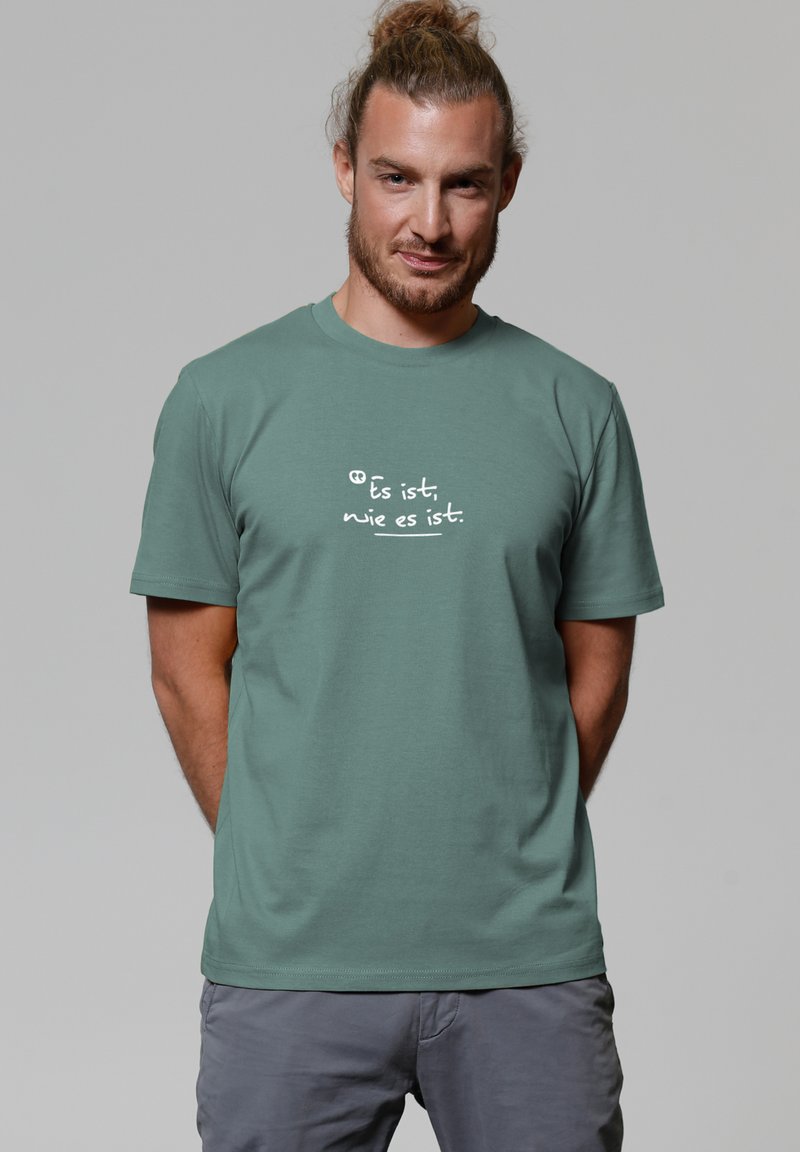 Mann mit hellbraunem Haar im Dutt, der ein grünes T-Shirt mit weißem deutschen Text "Es ist, wie es ist" und grauen Hosen vor einem grauen Hintergrund trägt.