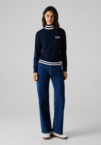 Maglione a collo alto blu con strisce bianche, logo sul petto, abbinato a jeans blu a gamba larga e scarpe chiare.