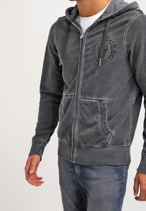 Hombre que lleva una sudadera con capucha gris oscuro desteñida con cremallera frontal y bolsillos, con un logo circular impreso en el pecho, combinado con vaqueros azules.