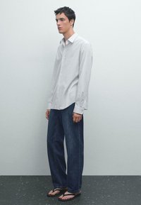  Lichtgrijs overhemd met lange mouwen, donkerblauwe jeans en zwarte slippers. Het overhemd heeft lange mouwen en de jeans hebben een relaxte pasvorm.