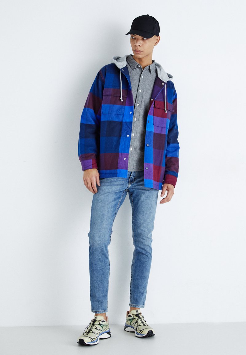 Helle, gefleckte Flanelljacke mit blauen, lila und schwarzen Karos, graue Kapuze und Knöpfe, kombiniert mit einem grauen Hemd, blauen Jeans und Turnschuhen.