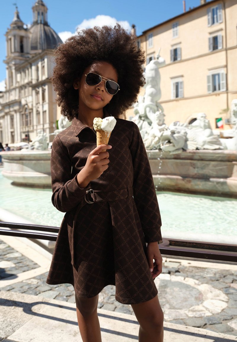 A girl in a dark brown, belted dress with a geometric pattern holds an ice cream cone with green and white scoops, wearing sunglasses.