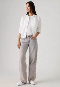 Ελαφρύ λευκό πουκάμισο με κουμπιά πάνω από ένα εφαρμοστό λευκό τοπ, συνδυασμένο με ψηλόμεσα, άνετα ανοιχτόχρωμα τζιν και λευκά αθλητικά παπούτσια.