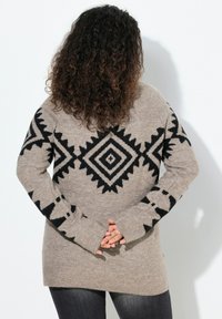 Beżowy długi sweter z czarnymi geometrycznymi wzorami, o szerokim dekolcie i długich rękawach. Miękka faktura i luźny krój.