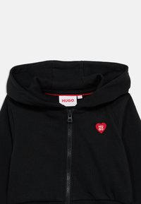 Μαύρο φούτερ με φερμουάρ σε απαλό ύφασμα, με κόκκινο κεντητό λογότυπο καρδιάς και επενδεδυμένη κουκούλα. Ορατό φερμουάρ και ριμπ τελείωμα στη μέση.