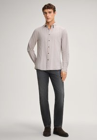 Camicia a maniche lunghe di colore grigio chiaro con righe verticali, colletto button-down e bottoni marroni, abbinata a jeans grigi scuri e stivali alla caviglia marroni.