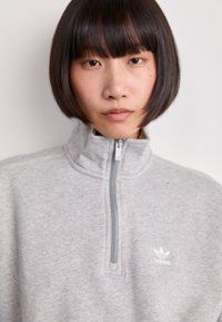 Sudadera con cremallera en color gris, hecha de una tela suave, con un cuello alto y un logo de Adidas bordado en blanco en el lado izquierdo del pecho.