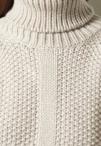 Dziany sweter z golfem w jasnokremowym kolorze z fakturowanym wzorem. Żebrowany dekolt i centralny panel dodają wizualnego uroku.