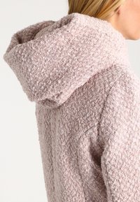 Lichtroze hoodie gemaakt van textuurstof. Heeft een dikke kraag en een ontspannen pasvorm met een subtiel geweven patroon.