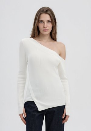 Giovane donna che indossa un maglione a coste bianco con una spalla scoperta e jeans scuri, in piedi davanti a uno sfondo grigio chiaro uniforme.