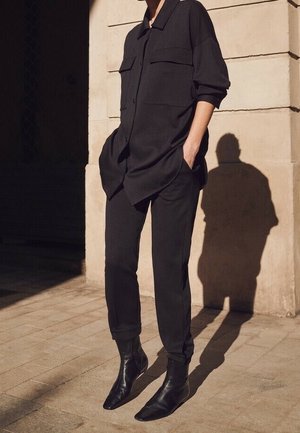 Person wearing loose black shirt with front pockets, black fitted pants, and pointed black ankle boots, standing with hands in pockets.