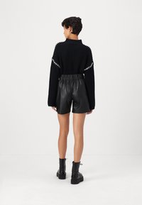 Svart stickad tröja med breda ärmar, parat med höga svarta shorts i konstläder. Modellen bär svarta ankelstövlar. Vita detaljer på ärmarna.