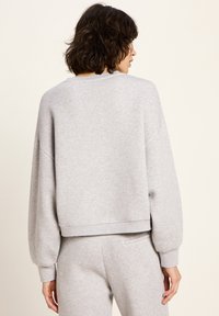 Eine Frau mit lockigem Haar trägt einen hellgrauen Langarmpullover und passende Hosen und steht mit dem Rücken zu einem schlichten, hellen Hintergrund.