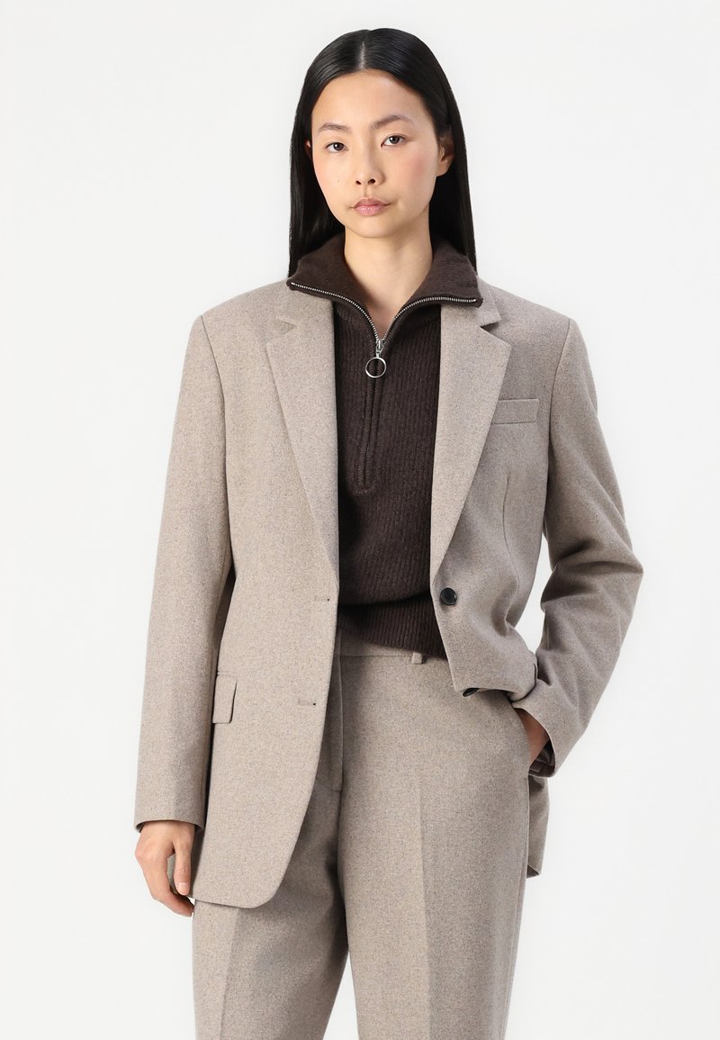 Giacca su misura marrone chiaro con spalle strutturate, bottone singolo e dettagli delle tasche frontali, abbinata a un maglione zip-up in maglia marrone scuro.
