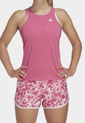 Woman wearing sleeveless pink athletic top and pink camo-patterned running shorts with hands on hips against a white background.