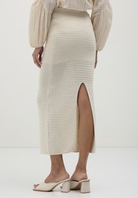 Jupon midi en tricot couleur crème avec des côtes verticales, fente sur le côté et finition texturée. Porté avec des mules à talons blocs beiges.