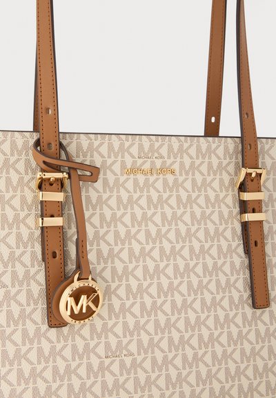 Sac cabas beige Michael Kors avec des sangles en cuir marron réglables et un pendentif logo MK doré sur une surface à motifs.