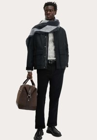 Schwarze Jacke, grauer Strickpullover, schwarze Hose und schwarze Schuhe. Grau und marineblauer karierter Schal umgehängt; braune Tasche mit Lederakzenten in der Hand.