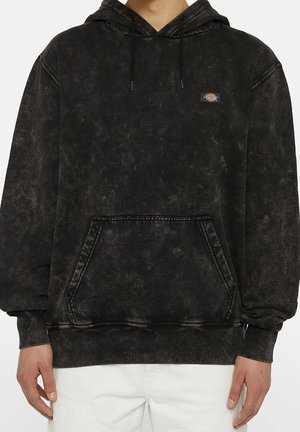 Czarna bluza z kapturem acid-wash z przednią kieszenią typu kangurek i małym wielokolorowym logo Dickies na piersi, noszona z białymi spodniami.