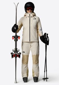 Attrezzatura da sci con un completo a due pezzi beige e crema con un accento bianco, guanti neri, casco nero e sci con attacchi rossi e bastoni.