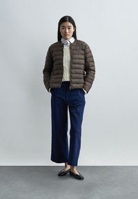 Γυναίκα που στέκεται με τα χέρια στις τσέπες, φορώντας καφέ μπουφάν με φουσκωτά μπαλώματα, εκρού πουλόβερ με πλέξη καλώδιο, μπλε παντελόνια με φαρδιές γάμπες και μαύρα μπαλαρίνες.