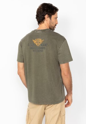 Homme aux cheveux bruns et à la barbe porte un t-shirt vintage vert olive avec l'inscription « Schott Air Crew » et un texte militaire, associé à un pantalon cargo beige.
