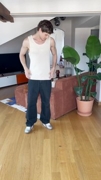 Weißes Tanktop, schwarze weit geschnittene Jeans, silberne Sneakers; steht auf einem Holzboden, im Hintergrund mit einer Pflanze in einem Topf und einer Couch.