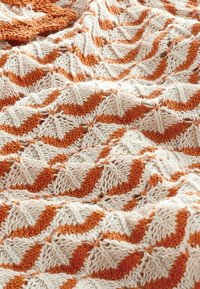 Gebreide deken met een gemêleerd zigzagpatroon in oranje en crème kleuren. Gemaakt van zachte garen, met afwisselende pieken en dalen.