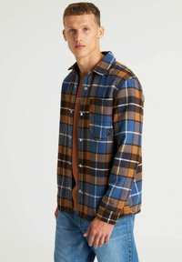 Flanellen overhemd in blauw, bruin en zwart geruit patroon; heeft drukknoopsluitingen, een borstzak en een relaxte pasvorm. Draagt over een bruin shirt.