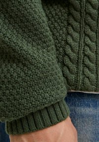 Donkergroene gebreide sweatermouw met geribde manchetten over een hand, met kabel- en textuursteekpatronen en daaronder blauwe denim jeans.