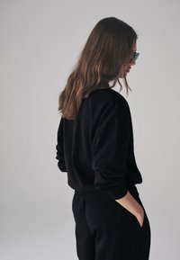 Mustast pehmest kangast lipsuga dressipluus, lõdva istuvusega, soonikkoes varrukate äärte ja küljetaskutega, sobitatud mustade pükstega.