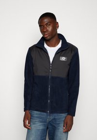 Veste en polaire marine avec un haut en nylon noir, fermeture éclaire intégrale, col haut et patch logo à l'avant, associée à un jean en denim bleu.