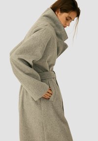 Szary wełniany płaszcz z wysokim kołnierzem, długimi rękawami i paskiem w talii. Posiada boczne kieszenie i miękką, teksturowaną tkaninę.