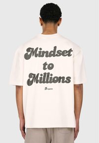 Lysecremet oversized t-shirt med fed sort tekst "Mindset to Millions" på ryggen, korte ærmer og en afslappet pasform.