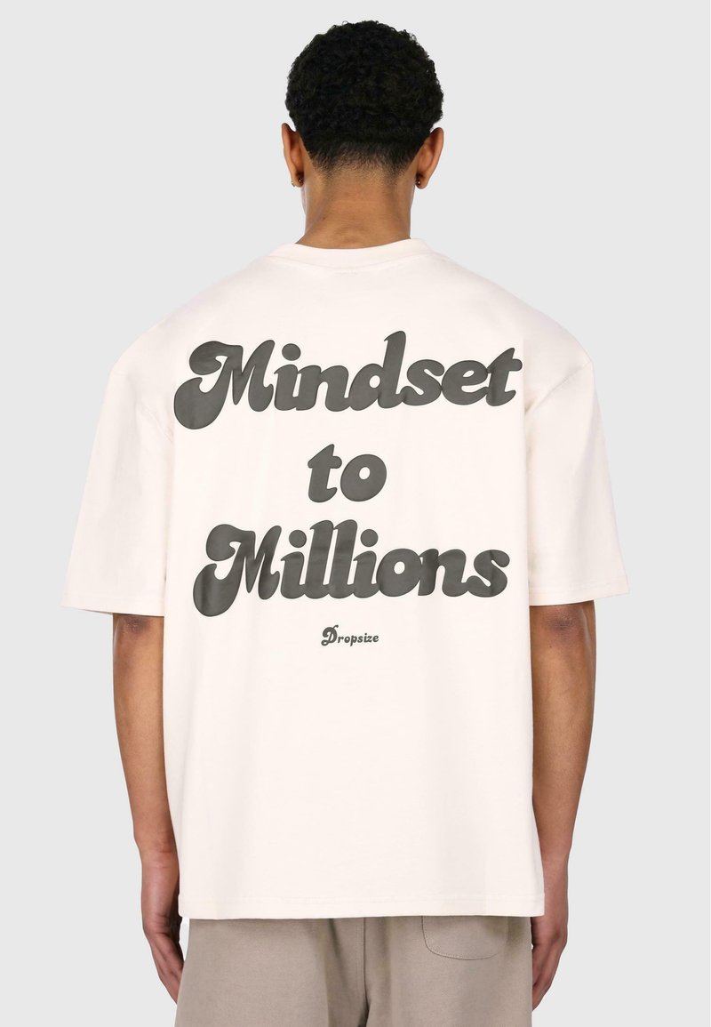 Lysecremet oversized t-shirt med fed sort tekst "Mindset to Millions" på ryggen, korte ærmer og en afslappet pasform.
