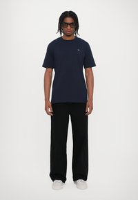 Mørkeblå bomulds t-shirt med korte ærmer og rund hals, parret med sorte brede bukser og hvide sneakers. Sorte solbriller bæres.
