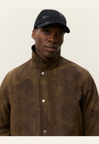 Braune, strukturierte Jacke mit hohem Kragen, Druckknöpfen und schwarzer Mütze mit Logo. Die Jacke hat eine gebürstete Optik.