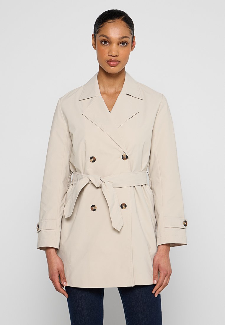 Trench beige con diseño de doble botonadura, cintura ajustada y cinturón. Presenta solapas clásicas y botones en los puños. Textura suave.