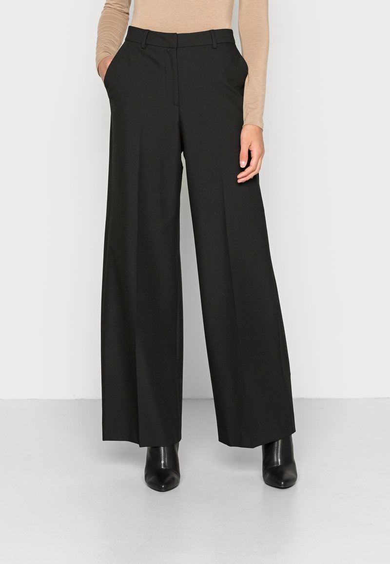 Kobieta w czarnych spodniaczkach typu wide-leg, beżowej bluzce z długim rękawem i czarnych botkach na obcasie, stoi z rękami w kieszeniach.