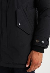 Svart vadderad jacka med strukturerad yta, utrustad med en stor lappficka och tryckknappar på ärmslutet för säker stängning och värme.