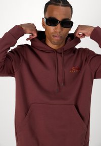 Burgundy kapucnis pulóver elülső zsebbel, húzózsinórral ellátott kapucnival és hímezett logóval. Sima textúra, laza szabás és hosszú ujjak.