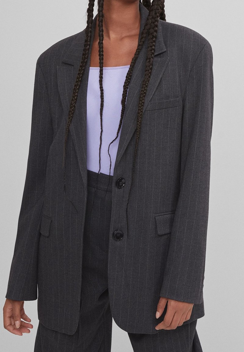 Personne portant un blazer gris foncé à fines rayures et un pantalon assorti sur un haut violet clair avec de longs cheveux tressés.