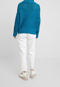 Pull turquoise en grosse maille avec un col montant, associé à un pantalon blanc raccourci et des chaussures à plateforme beiges, mettant en valeur un tissu texturé.