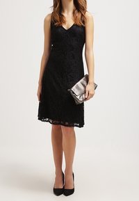 Robe en dentelle noire avec un décolleté en V, une silhouette ajustée et une longueur de genou. Assortie à une pochette argentée et des escarpins noirs.