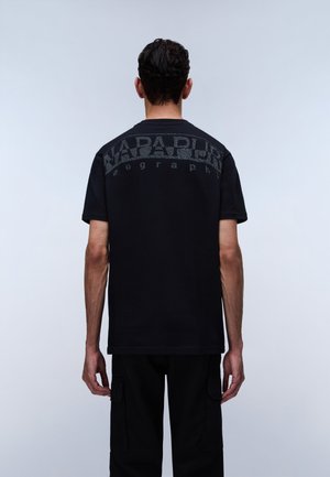 Homme de dos portant un t-shirt noir à manches courtes avec le texte effacé "Napapijri" sur le haut du dos, et un pantalon cargo noir.